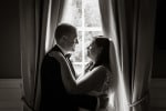 A black and white photo of a wedding couple framed in the curtains by a window