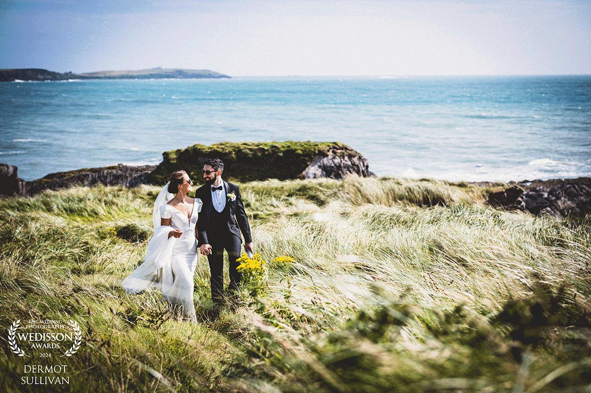 bride-and-groom-cliff-sea-sky A bride and groom walk hand in hand along a clifftop with the sea behind them - Cork Wedding Photographs