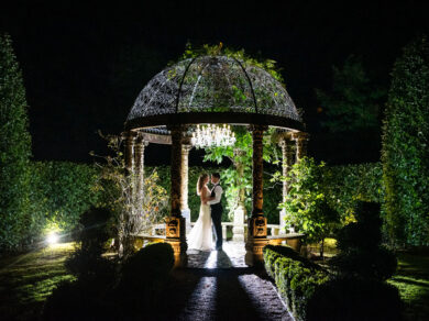 A bride and groom stand backlit at night by off camera flash in the temple in the gardens of Ballyseede Castle in Tralee, Ireland