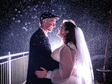 A backlit bride and groom embrace in the rain at night with a white railing beside them